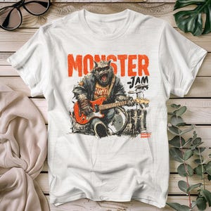 May include: White t-shirt with a graphic of a monster playing a red electric guitar, with the words "MONSTER JAM" in orange. The monster is wearing a jacket and is playing drums. The shirt has a vintage, rock-and-roll aesthetic.