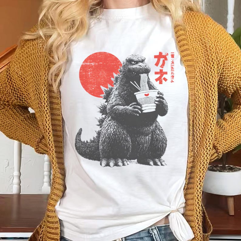 Puede incluir: Camiseta blanca con un gr&aacute;fico de Godzilla comiendo fideos ramen, con un sol rojo de fondo. Texto japon&eacute;s impreso en el lado derecho del gr&aacute;fico. La camiseta es una prenda informal.
