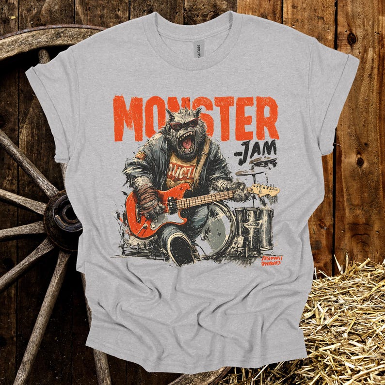 May include: A light gray t-shirt featuring a monster playing a red electric guitar in front of a drum set. The text "MONSTER JAM" is printed above the monster. The shirt has rolled-up sleeves.