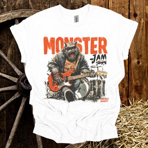 May include: White t-shirt with a graphic of a monster playing a red guitar and drums. The text "MONSTER JAM" is printed in orange above the monster. The shirt has a crew neck and short sleeves.