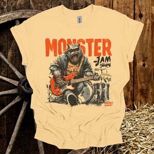 May include: A light yellow t-shirt with a graphic of a monster playing a red guitar and drums. The text "MONSTER JAM" is printed above the monster. The shirt is made of a soft material.