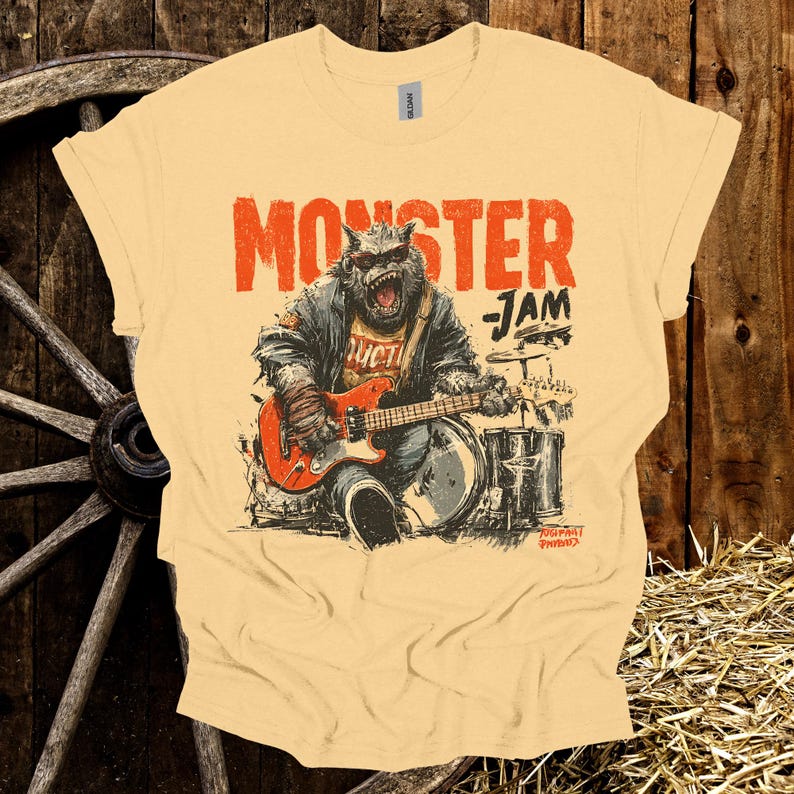 May include: A light yellow t-shirt with a graphic of a monster playing a red guitar and drums. The text "MONSTER JAM" is printed above the monster. The shirt is made of a soft material.