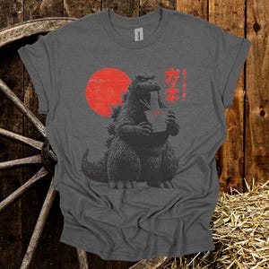 Puede incluir: Camiseta gris oscuro con un dise&ntilde;o de Godzilla comiendo fideos, con un sol rojo de fondo. Texto japon&eacute;s impreso a la derecha. La camiseta est&aacute; hecha de un material suave.