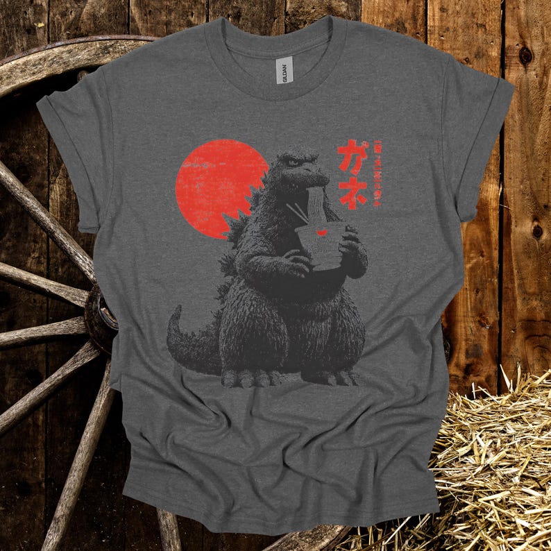 Puede incluir: Camiseta gris oscuro con un dise&ntilde;o de Godzilla comiendo fideos, con un sol rojo de fondo. Texto japon&eacute;s impreso a la derecha. La camiseta est&aacute; hecha de un material suave.
