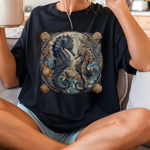 May include: Navy blue t-shirt featuring a detailed circular design with two seahorses, seashells, and ocean waves in shades of blue, beige, and gold. The design is centered on the front of the shirt.