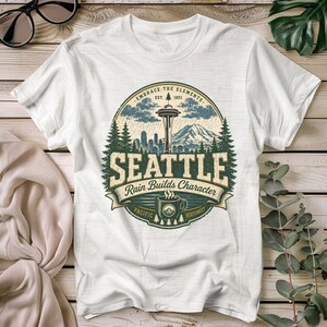 May include: A white short-sleeve t-shirt featuring a circular graphic of the Seattle skyline, including the Space Needle, pine trees, and a mountain range. The text reads EMBRACE THE ELEMENTS, EST. 1851, SEATTLE, Rain Builds Character, and PACIFIC NORTHWEST.