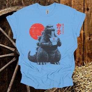 Puede incluir: Camiseta azul claro con un dise&ntilde;o de Godzilla comiendo ramen, con un sol rojo de fondo. Texto japon&eacute;s encima del dise&ntilde;o. La camiseta est&aacute; hecha de un material suave.
