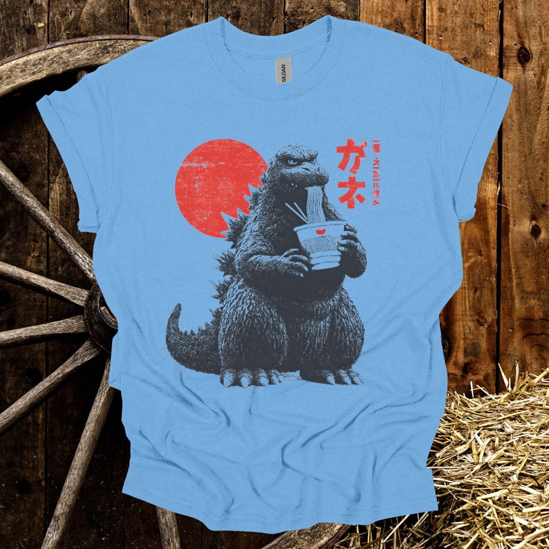 Puede incluir: Camiseta azul claro con un dise&ntilde;o de Godzilla comiendo ramen, con un sol rojo de fondo. Texto japon&eacute;s encima del dise&ntilde;o. La camiseta est&aacute; hecha de un material suave.