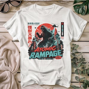 Op de afbeelding: Wit T-shirt met een Godzilla-afbeelding. Het ontwerp bevat een rode zon, een Godzilla-silhouet en de tekst "ATOMIC RAMPAGE" in turquoise en rood. Japanse tekst is ook aanwezig.
