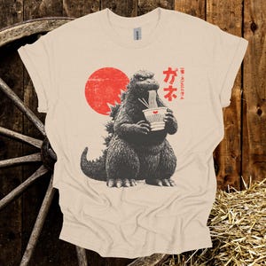 Puede incluir: Camiseta beige con un dise&ntilde;o de Godzilla comiendo ramen con un sol rojo de fondo. Texto japon&eacute;s encima del dise&ntilde;o de Godzilla. La camiseta est&aacute; hecha de un material suave.