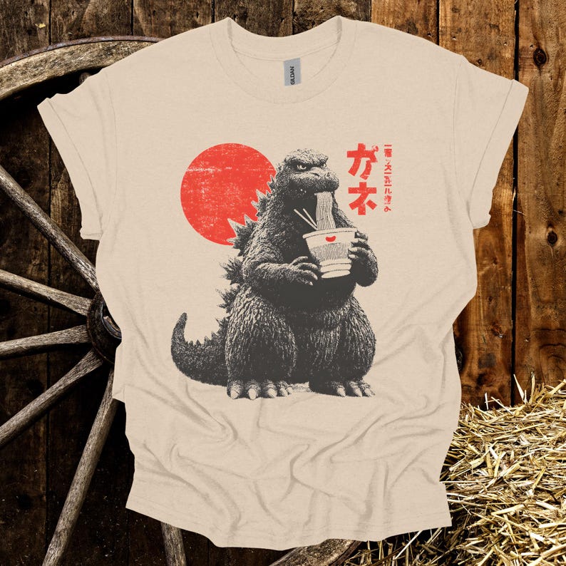 Puede incluir: Camiseta beige con un dise&ntilde;o de Godzilla comiendo ramen con un sol rojo de fondo. Texto japon&eacute;s encima del dise&ntilde;o de Godzilla. La camiseta est&aacute; hecha de un material suave.