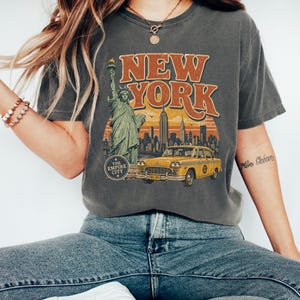 May include: Dark grey t-shirt with a vintage New York City design. The graphic shows the Statue of Liberty, a yellow taxi, and the words "NEW YORK" in orange. The Empire State Building is also depicted.