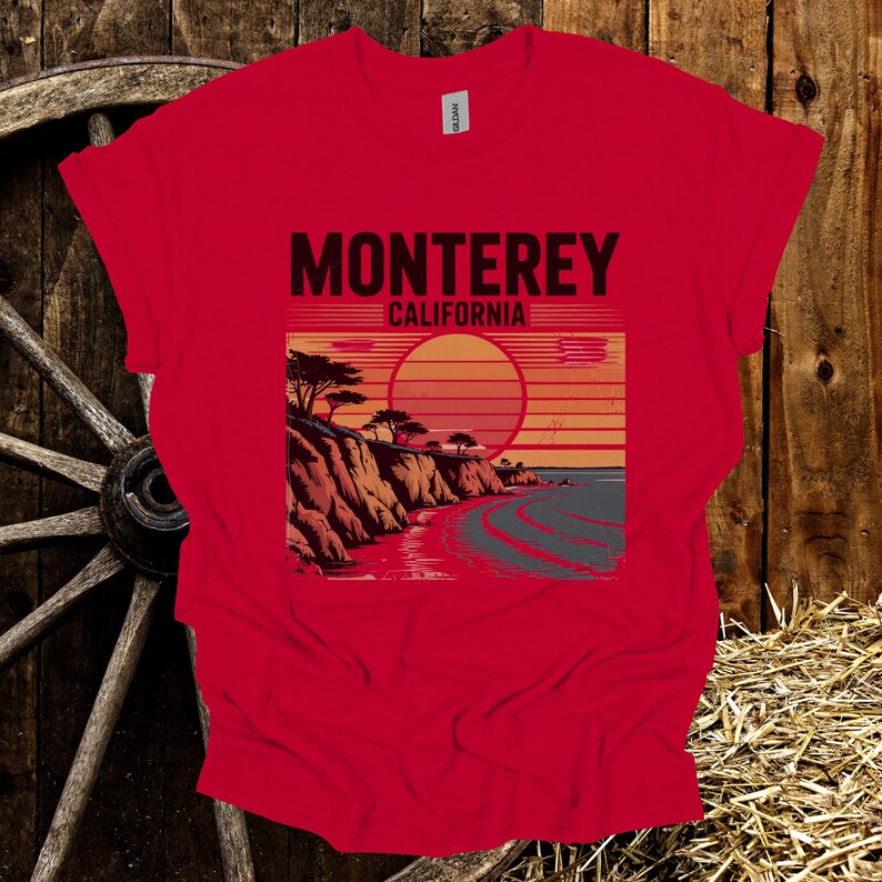 May include: Red t-shirt featuring a graphic of the California coastline with the text "Monterey California". The design includes a sunset, cliffs, and ocean waves. The shirt is made of a soft material.