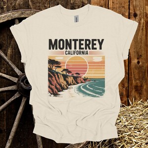 May include: Cream-colored t-shirt featuring a graphic of a coastal landscape with the text "MONTEREY CALIFORNIA". The design includes a sunset, cliffs, trees, and ocean waves. The shirt is made of a soft material.