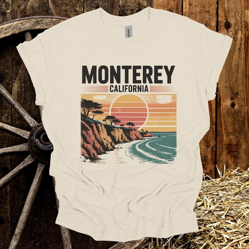 May include: Cream-colored t-shirt featuring a graphic of a coastal landscape with the text "MONTEREY CALIFORNIA". The design includes a sunset, cliffs, trees, and ocean waves. The shirt is made of a soft material.