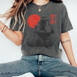 Puede incluir: Camiseta gris oscuro con un dise&ntilde;o de Godzilla comiendo ramen, con un sol rojo de fondo. Texto japon&eacute;s encima del dise&ntilde;o. La camiseta es de estilo casual y c&oacute;modo.