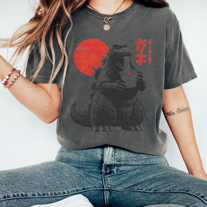Puede incluir: Camiseta gris oscuro con un dise&ntilde;o de Godzilla comiendo ramen, con un sol rojo de fondo. Texto japon&eacute;s encima del dise&ntilde;o. La camiseta es de estilo casual y c&oacute;modo.