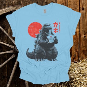 Puede incluir: Camiseta azul claro con un dise&ntilde;o de Godzilla comiendo fideos ramen. El dise&ntilde;o incluye un sol rojo y texto japon&eacute;s. El gr&aacute;fico est&aacute; en tonos grises y negros.