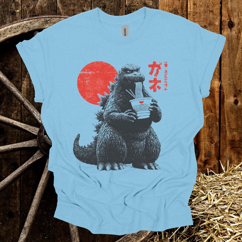 Puede incluir: Camiseta azul claro con un dise&ntilde;o de Godzilla comiendo fideos ramen. El dise&ntilde;o incluye un sol rojo y texto japon&eacute;s. El gr&aacute;fico est&aacute; en tonos grises y negros.