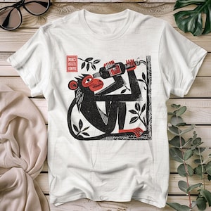 Puede incluir: Camiseta blanca con un diseño en negro y rojo de un mono sosteniendo una botella de vino. El mono está representado en un estilo de grabado en madera, con el texto "MIKS MARVIN JONYE" en un recuadro rojo. La camiseta tiene cuello redondo.