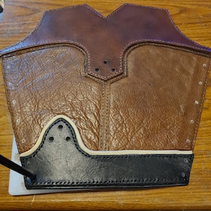 May include: A brown leather arm guard with a textured surface. The guard has a black leather trim at the bottom and a white accent line. The top portion is a darker brown. There are several small holes and stitching details.