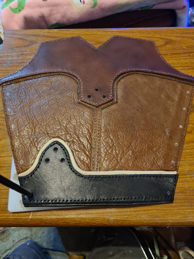 May include: A brown leather arm guard with a textured surface. The guard has a black leather trim at the bottom and a white accent line. The top portion is a darker brown. There are several small holes and stitching details.