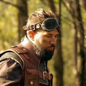 May include: A brown leather vest with multiple pockets and a matching leather strap. The person is wearing vintage-style goggles with a brown leather strap. The background is a blurred forest.