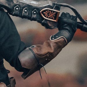May include: Close-up of a black and brown leather arm guard and glove, with a decorative metal piece featuring a cross design. The arm guard is brown with black accents and a leather tie.