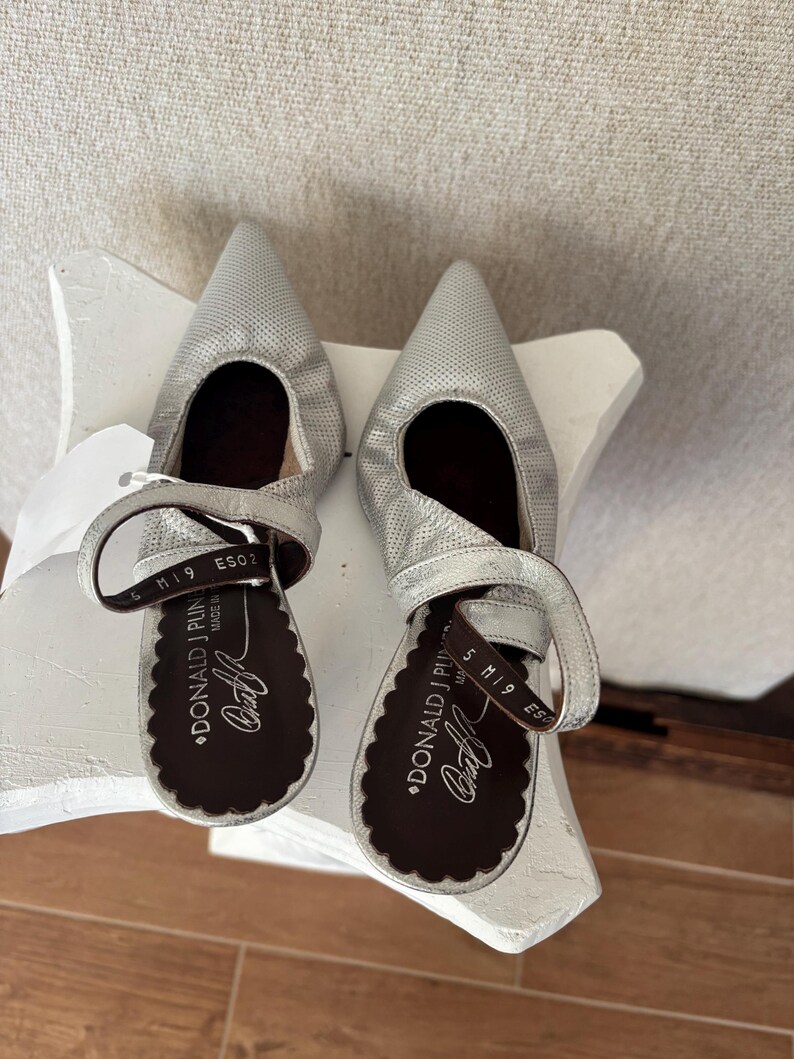 Puede incluir: Un par de zapatos de sal&oacute;n plateados de cuero perforado con punta y correas en el tobillo. Los zapatos tienen una suela festoneada y la marca "DONALD J PLIN" es visible en la plantilla. Los tacones miden aproximadamente 5 cm.