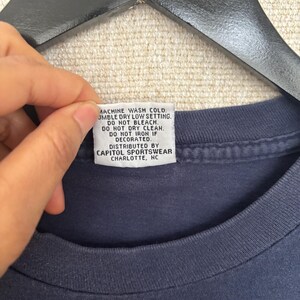May include: A navy blue t-shirt with a tag that reads "MACHINE WASH COLD. TUMBLE DRY LOW SETTING. DO NOT BLEACH. DO NOT DRY CLEAN. DO NOT IRON IF DECORATED. DISTRIBUTED BY CAPITOL SPORTSWEAR CHARLOTTE, NC."