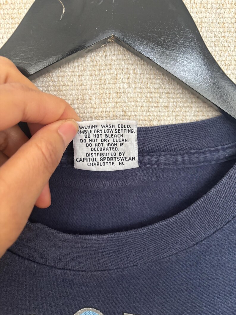 May include: A navy blue t-shirt with a tag that reads "MACHINE WASH COLD. TUMBLE DRY LOW SETTING. DO NOT BLEACH. DO NOT DRY CLEAN. DO NOT IRON IF DECORATED. DISTRIBUTED BY CAPITOL SPORTSWEAR CHARLOTTE, NC."