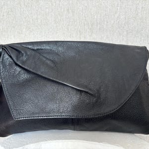 May include: A black leather clutch with a textured surface. The clutch has a flap closure and a knotted detail on the side. The bag is designed to be carried by hand.