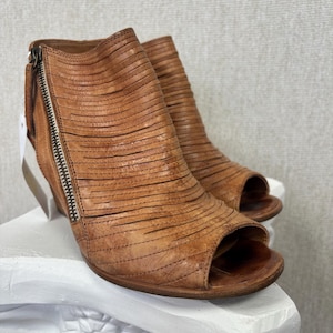 Puede incluir: Botines peep-toe de cuero marrón con tacón de cuña. Los zapatos tienen una cremallera lateral y un diseño de flecos en capas. Se exhiben sobre un pedestal blanco. El tacón mide aproximadamente 7.6 cm.