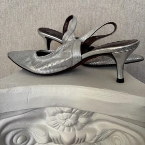 Puede incluir: Zapatos de tac&oacute;n plateados con correa trasera, punta afilada y tac&oacute;n peque&ntilde;o y curvado. Los zapatos tienen un dise&ntilde;o perforado en la parte superior y una correa que rodea el tobillo. Los tacones miden unos 6,35 cm de alto.