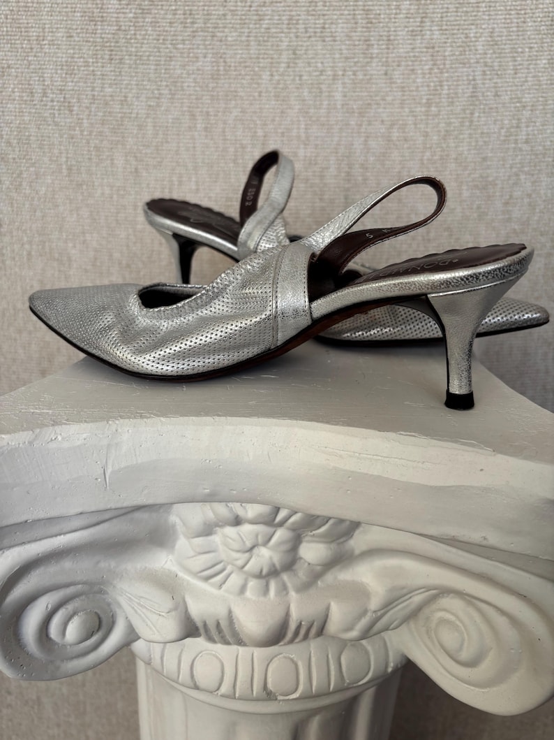 Puede incluir: Zapatos de tac&oacute;n plateados con correa trasera, punta afilada y tac&oacute;n peque&ntilde;o y curvado. Los zapatos tienen un dise&ntilde;o perforado en la parte superior y una correa que rodea el tobillo. Los tacones miden unos 6,35 cm de alto.