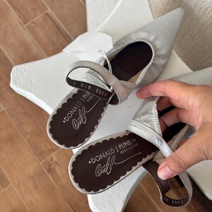 Puede incluir: Bailarinas plateadas de punta con correa. Los zapatos tienen un aspecto texturizado y una suela festoneada. La marca "DONALD J PLINER" es visible en la plantilla, con "MADE IN ITALY" debajo.