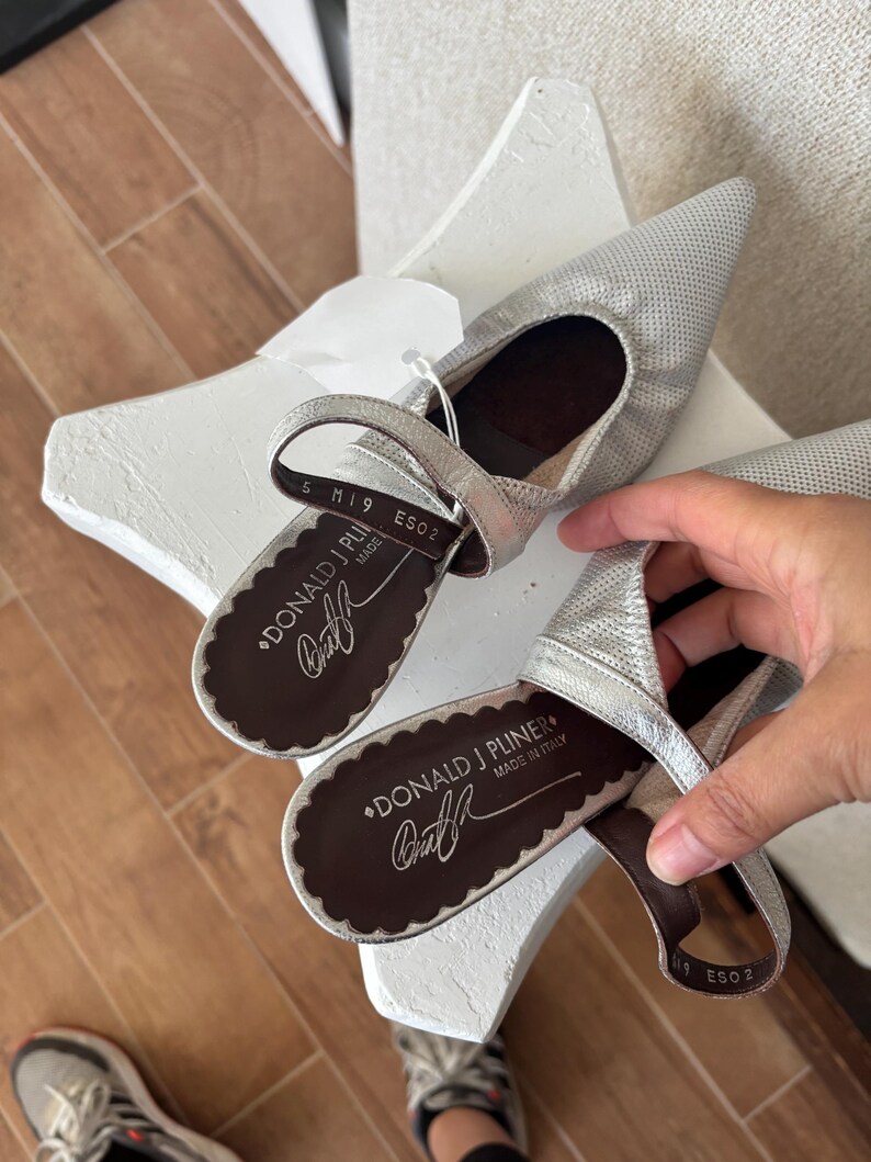 Puede incluir: Bailarinas plateadas de punta con correa. Los zapatos tienen un aspecto texturizado y una suela festoneada. La marca "DONALD J PLINER" es visible en la plantilla, con "MADE IN ITALY" debajo.