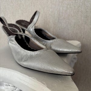 Puede incluir: Un par de mules de cuero plateado perforado con punta. Los zapatos tienen un tac&oacute;n bajo y una correa trasera. El interior del zapato es marr&oacute;n oscuro. Los zapatos se exhiben sobre un pedestal decorativo blanco.