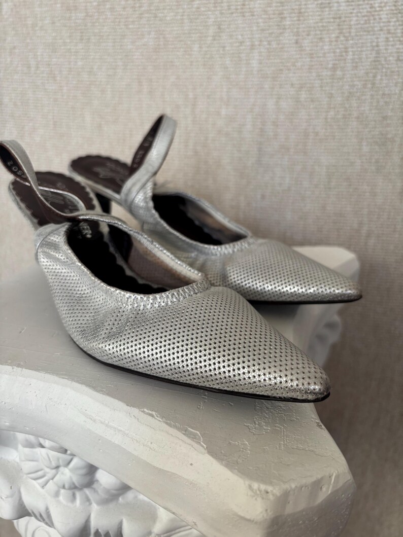 Puede incluir: Un par de mules de cuero plateado perforado con punta. Los zapatos tienen un tac&oacute;n bajo y una correa trasera. El interior del zapato es marr&oacute;n oscuro. Los zapatos se exhiben sobre un pedestal decorativo blanco.