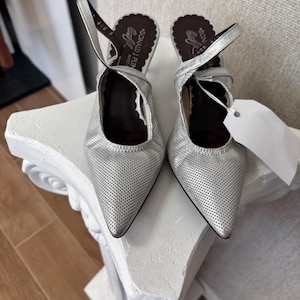 Puede incluir: Un par de tacones de cuero plateado perforado con punta y correas en el tobillo. Los tacones tienen un ribete festoneado y la marca "DONALD J PLINER" es visible en la plantilla. Una etiqueta blanca cuelga de un zapato.