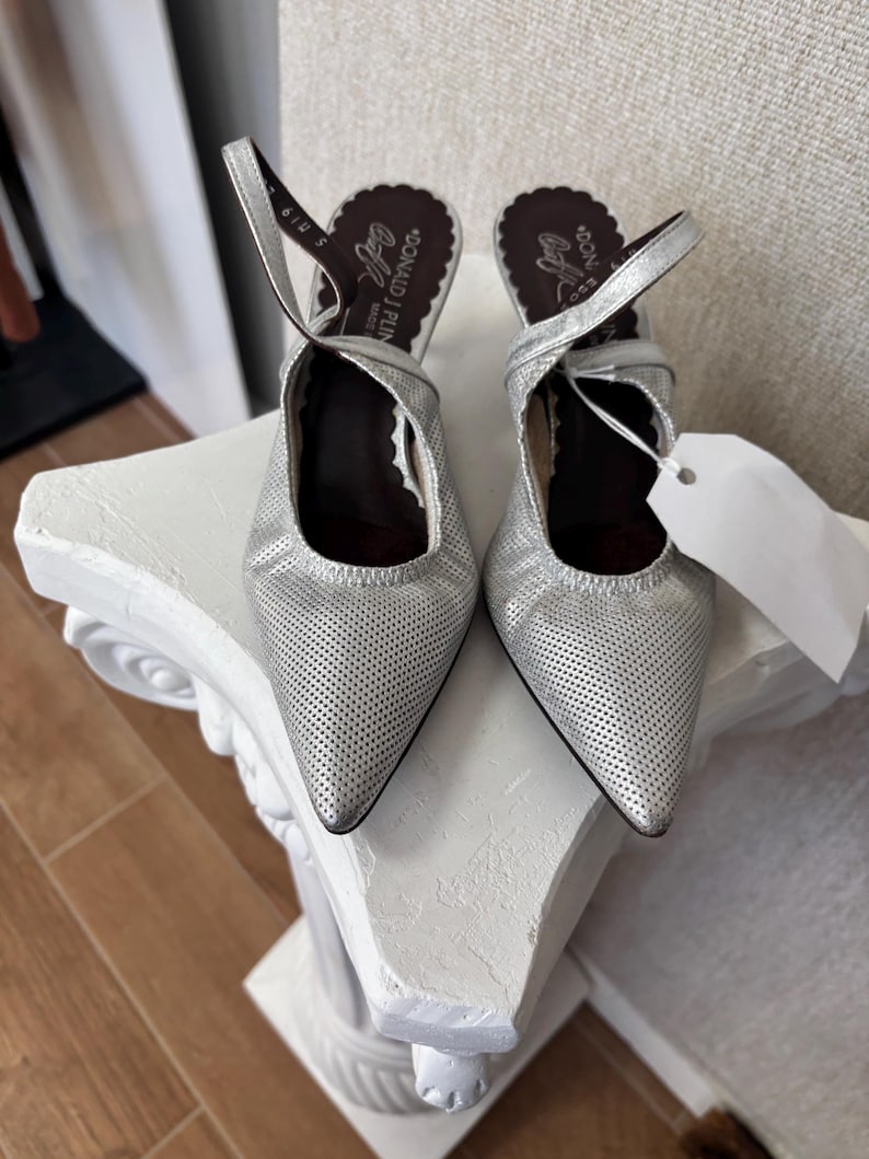 Puede incluir: Un par de tacones de cuero plateado perforado con punta y correas en el tobillo. Los tacones tienen un ribete festoneado y la marca "DONALD J PLINER" es visible en la plantilla. Una etiqueta blanca cuelga de un zapato.
