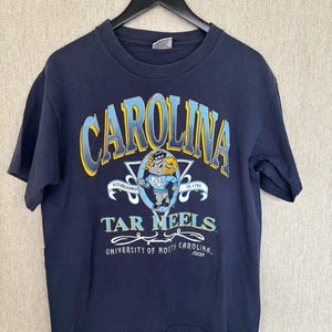 May include: Navy blue t-shirt featuring the word "CAROLINA" in light blue and yellow arched lettering. Below the word "CAROLINA" is the text "TAR HEELS" and a graphic of a ram. The shirt also includes the text "UNIVERSITY OF NORTH CAROLINA."