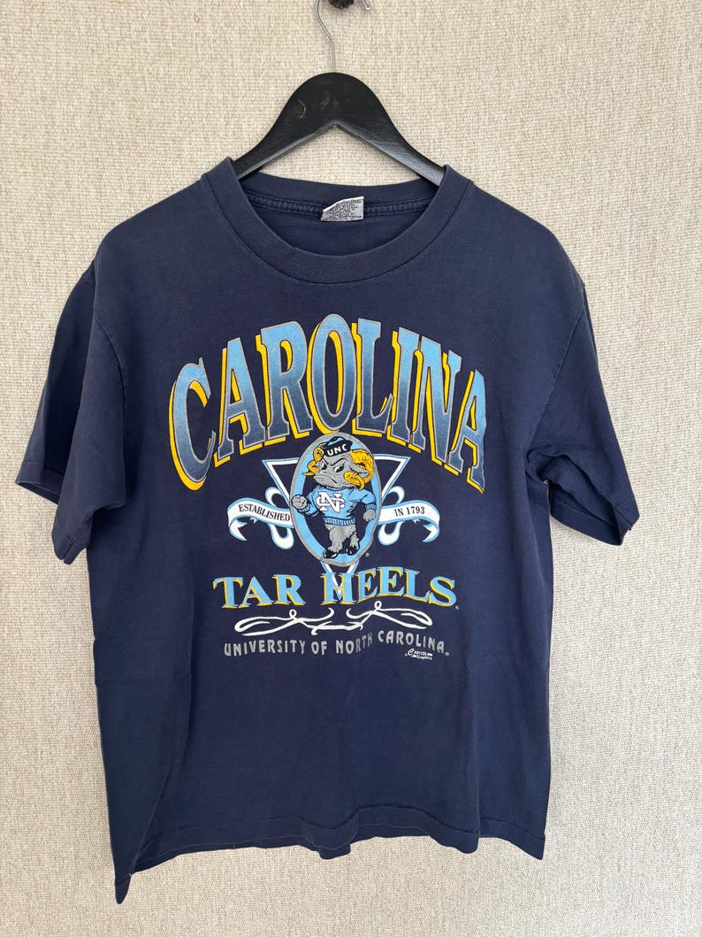 May include: Navy blue t-shirt featuring the word "CAROLINA" in light blue and yellow arched lettering. Below the word "CAROLINA" is the text "TAR HEELS" and a graphic of a ram. The shirt also includes the text "UNIVERSITY OF NORTH CAROLINA."