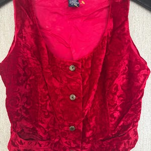 May include: A red velvet vest with a floral pattern and three decorative buttons. The vest has a scoop neckline and two front pockets. The GAP label is visible at the top of the vest.