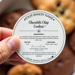 May include: A round label for chocolate chip cookies, held by a hand, with the text "YOUR BAKERY NAME" and "Chocolate Chip Cookies." The label lists ingredients, net weight 227g, and allergen information. The label also includes the bakery's name and address.