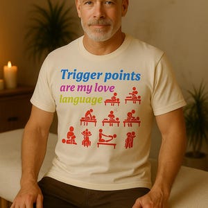 Puede incluir: Una camiseta color crema con el texto "Trigger points are my love language" en azul y verde. Ilustraciones rojas de técnicas de masaje están debajo del texto. La persona lleva pantalones marrones.