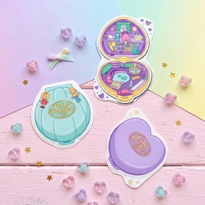 May include: Three Polly Pocket stickers featuring a teal shell-shaped case with two figures, an open heart-shaped case revealing a detailed interior room and garden scene, and a closed purple heart-shaped case, all decorated with small heart accents.