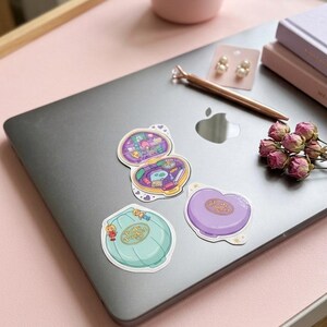 May include: Three vinyl stickers featuring Polly Pocket designs. One sticker shows an open compact with a detailed interior, while the other two depict closed compacts in light blue and purple, each labeled with the Polly Pocket brand name.