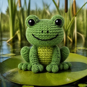 Peut inclure: Une grenouille verte au crochet faite à la main, avec de grands yeux noirs et une bouche souriante. La grenouille est assise sur une feuille de nénuphar verte dans un étang. Le ventre de la grenouille est vert clair. L'arrière-plan montre des roseaux et de l'eau.