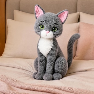 May include: A gray and white plush cat with pink inner ears and green eyes sits upright on a bed. The handmade cat has a white bib and a long tail. The cat is a stuffed animal.